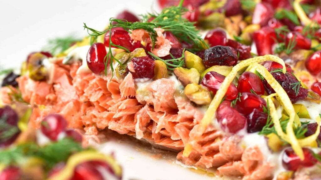Close-up of cooked salmon topped with pomegranate seeds, pistachios, lemon zest, dill, and a creamy white sauce.