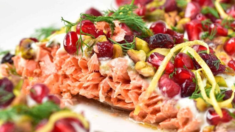 Close-up of cooked salmon topped with pomegranate seeds, pistachios, lemon zest, dill, and a creamy white sauce.