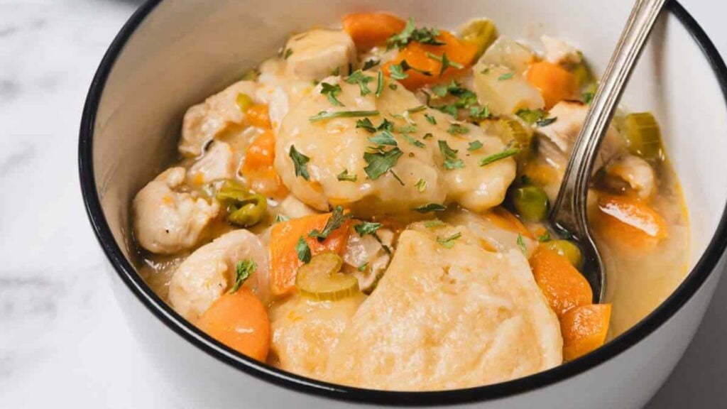 A bowl of chicken and dumplings with carrots, celery, and parsley, served with a spoon.
