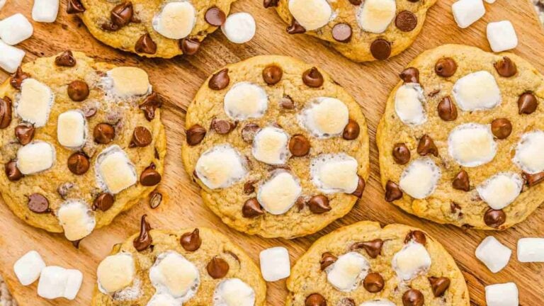 Chocolate chip cookies topped with mini marshmallows and chocolate chips, arranged on a wooden surface with extra marshmallows scattered around.