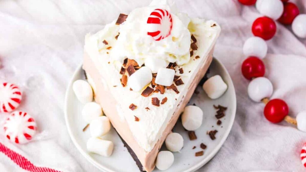A slice of whipped cream-topped pie with chocolate shavings, mini marshmallows, and a peppermint candy on a small plate, surrounded by more marshmallows—one of those easy desserts perfect for your New Year’s Eve desserts table.