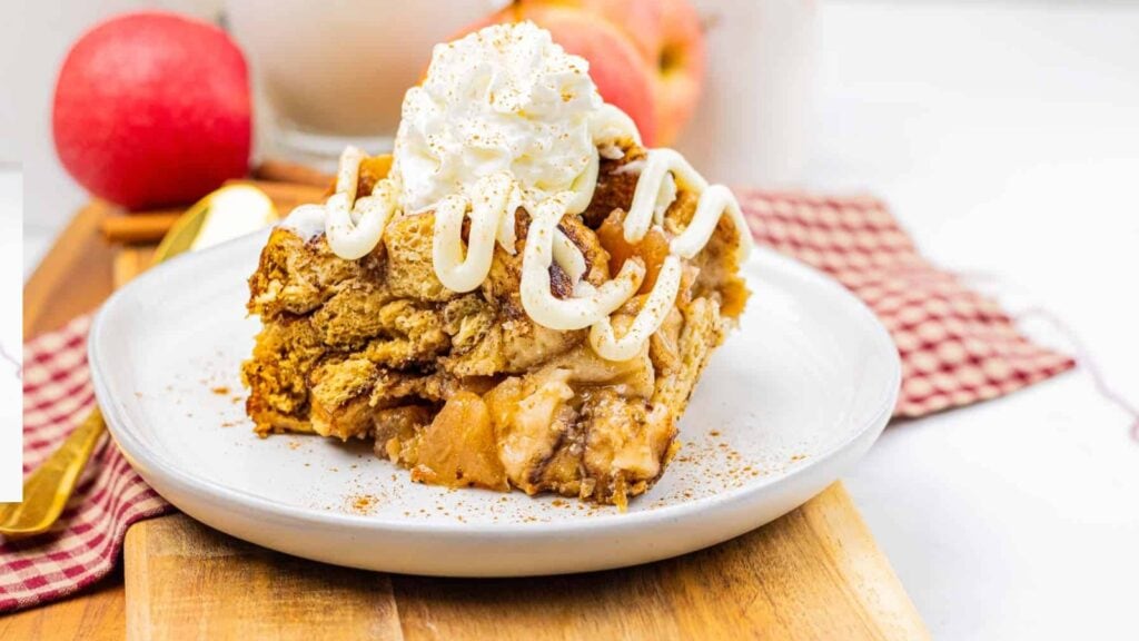 A slice of apple pie topped with whipped cream and icing sits on a white plate, with apples and a red-checkered napkin in the background.