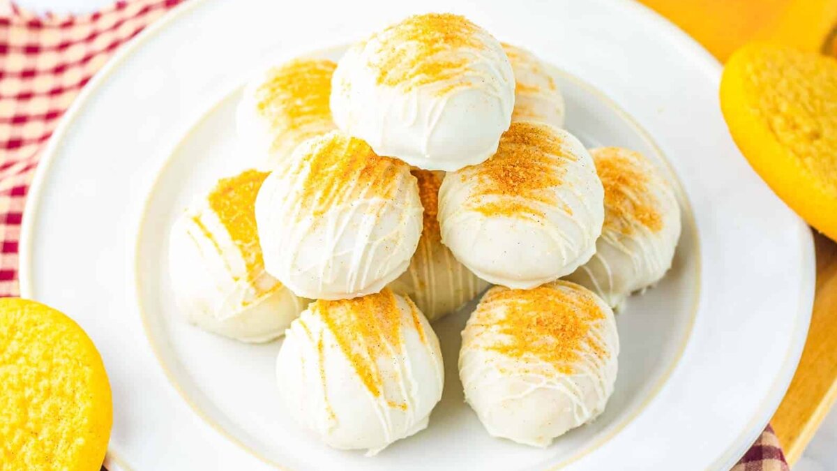 A plate of round, white chocolate-coated dessert balls topped with golden crumbs, arranged in a pyramid shape. Nearby, there are yellow cookies and a red checkered cloth.