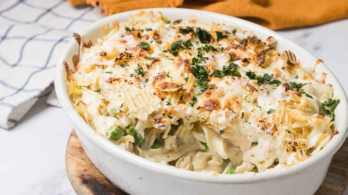 A fork lifts creamy pasta bake with peas and cheese from a casserole dish, garnished with herbs.