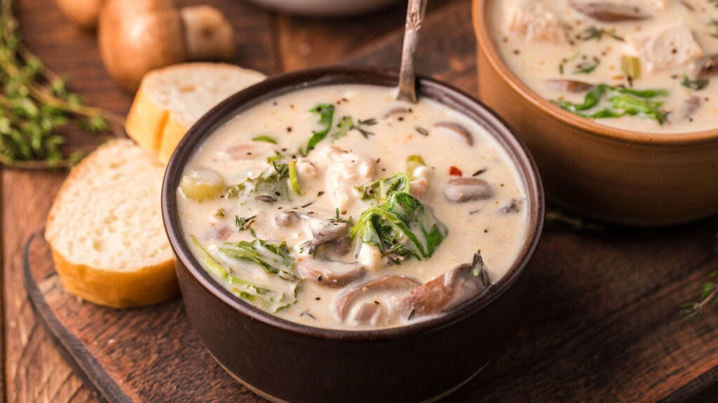 A bowl of creamy soup with mushrooms, leafy greens, and herbs sits on a wooden surface next to slices of bread.