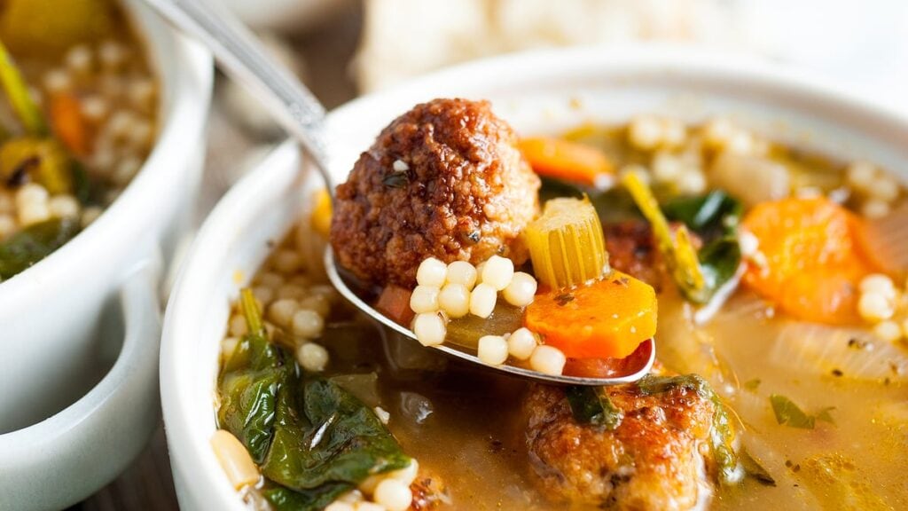A close-up of a bowl of soup with meatballs, carrots, celery, spinach, and pearl pasta, with a spoon lifting out a meatball and vegetables.