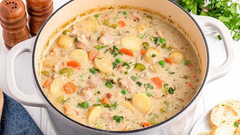 A pot of creamy chicken stew with sliced potatoes, carrots, peas, and herbs on a light-colored tabletop with parsley and salt and pepper shakers nearby.