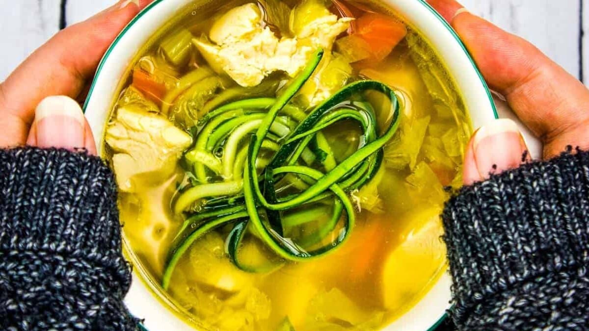 A person holding a bowl of chicken and zucchini soup.