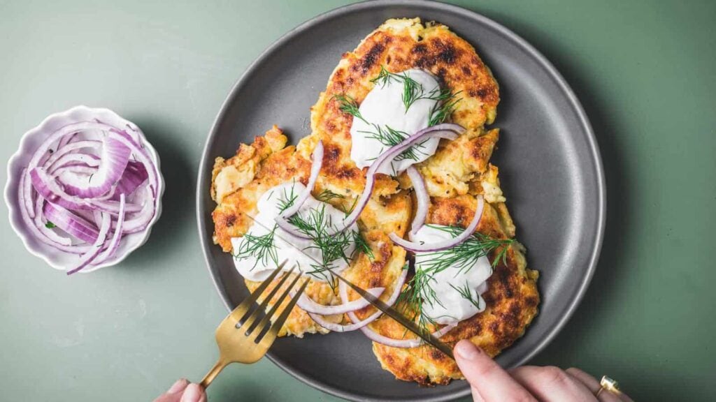 Three potato pancakes topped with sour cream, red onion slices, and dill on a gray plate, with a fork and knife cutting into one pancake. A small bowl of red onion slices is beside the plate.