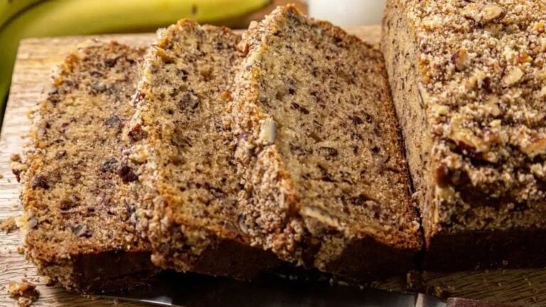 Three slices of banana bread with a crumbly topping rest on a wooden cutting board, with a loaf and part of a banana visible in the background.