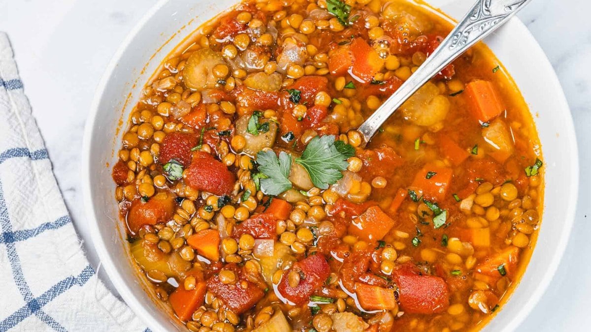 A bowl of lentil soup with carrots, tomatoes, herbs, and a spoon, garnished with parsley on top.
