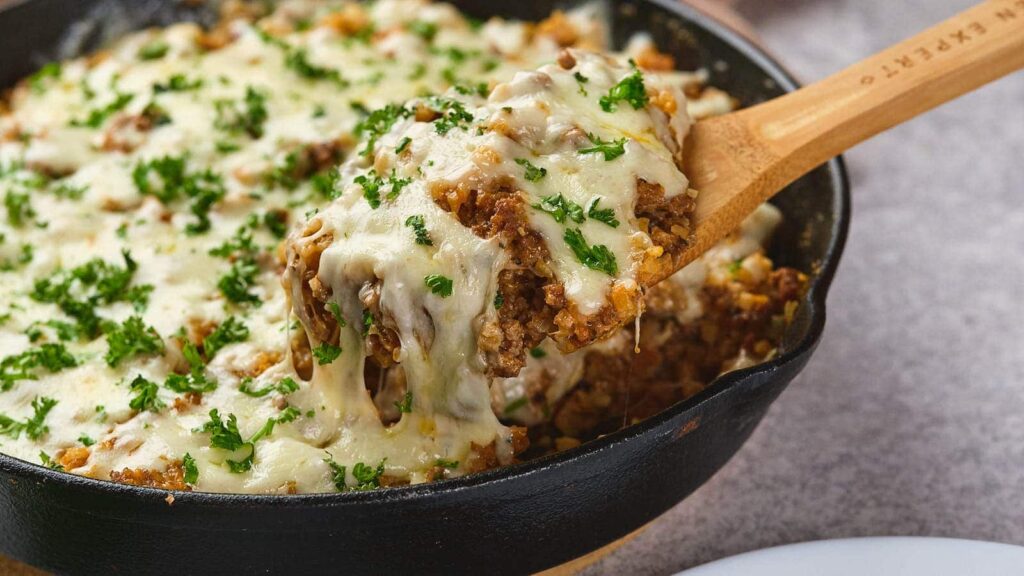 A wooden spoon lifts a portion of cheesy ground meat casserole topped with melted cheese and chopped parsley from a black cast iron skillet.
