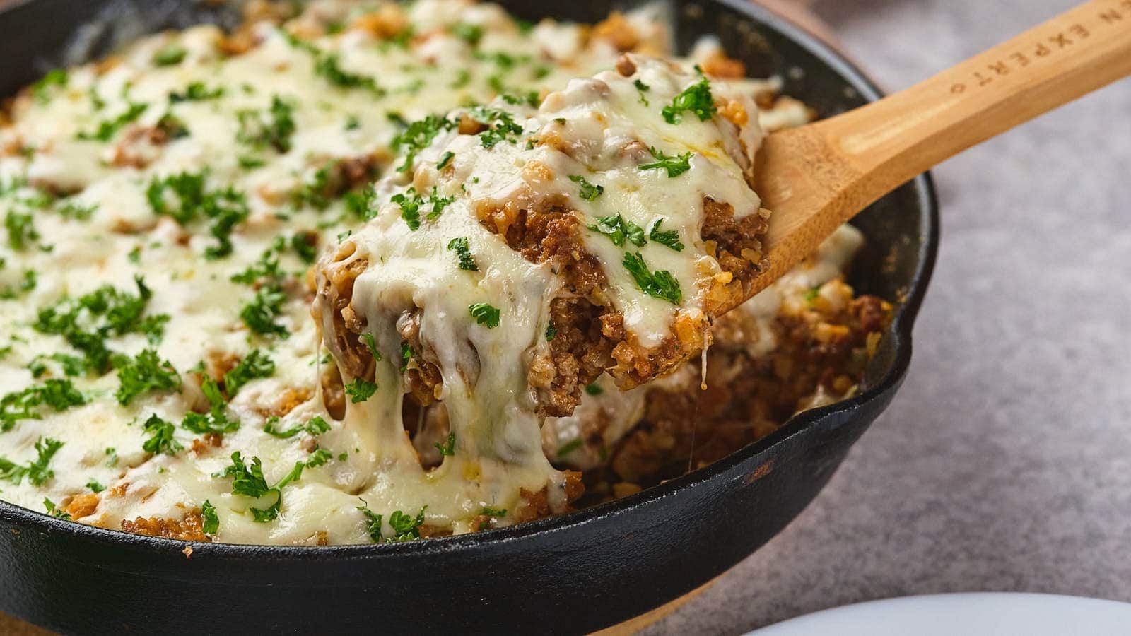 A wooden spoon lifts a portion of cheesy ground meat casserole topped with melted cheese and chopped parsley from a black cast iron skillet.