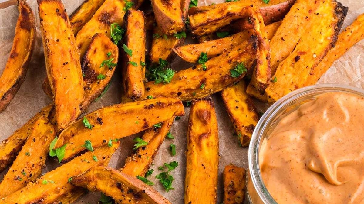 Sweet potato fries garnished with chopped parsley are arranged on parchment paper next to a small bowl of creamy dipping sauce.