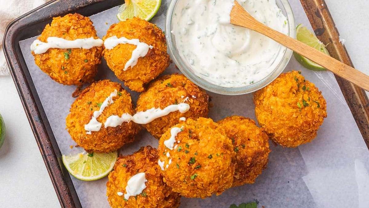 Seven golden-brown fritters topped with white sauce are arranged on parchment next to lime wedges and a bowl of creamy dip with a wooden spoon.