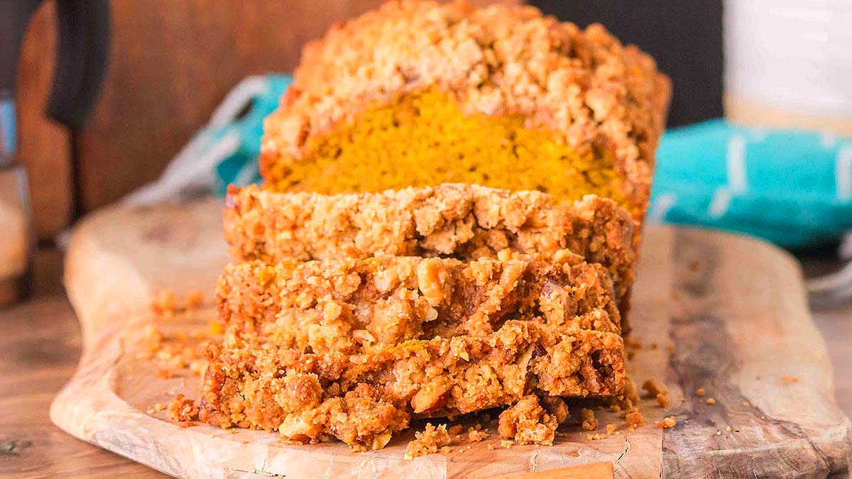 A loaf of crumb-topped pumpkin bread sits on a wooden cutting board with several slices cut and fanned out in front.