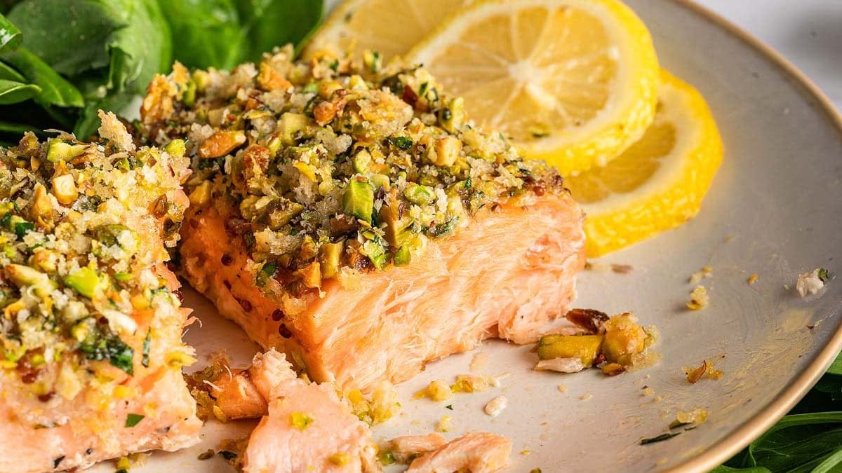 A plate with two slices of pistachio-crusted salmon, garnished with lemon slices and spinach leaves.