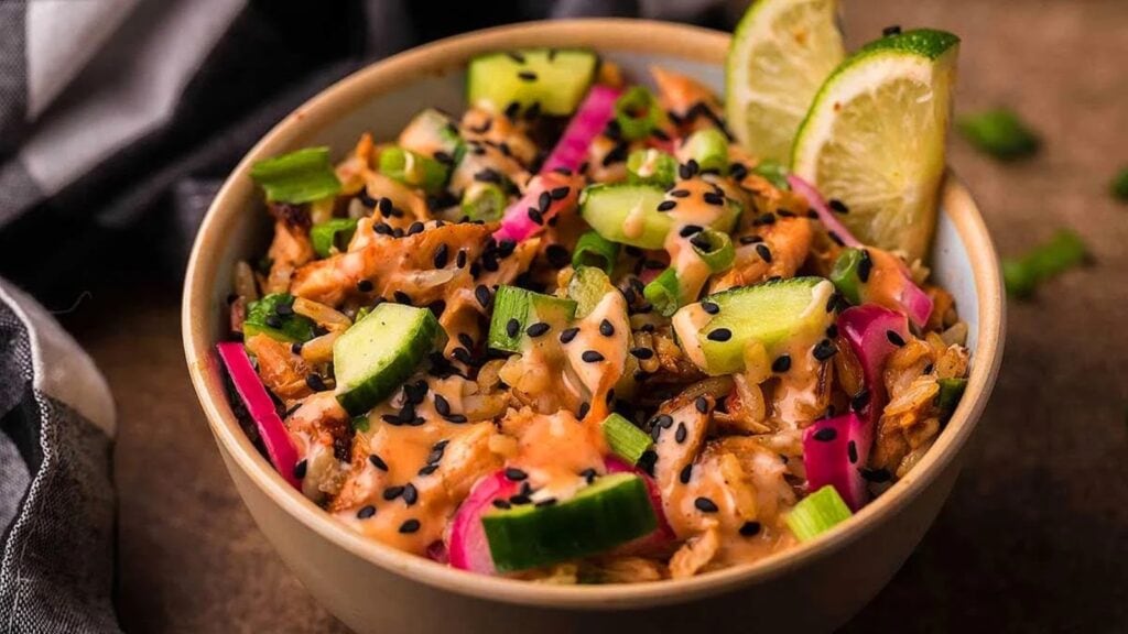 A bowl filled with a mixed salad containing diced cucumber, pickled onions, shredded meat, green onions, black sesame seeds, and lime wedges.