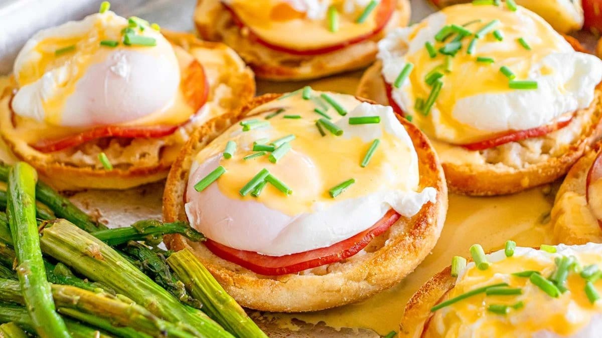 Eggs Benedict with poached eggs, ham, and hollandaise sauce on English muffins, garnished with chives, served with roasted asparagus.