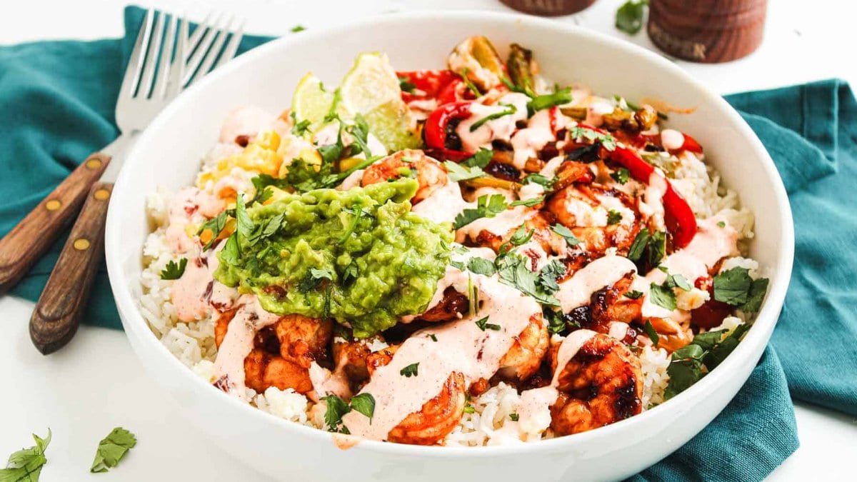 A white bowl filled with rice, grilled chicken, guacamole, corn, peppers, cilantro, and a creamy sauce, placed on a green napkin with two forks beside it.