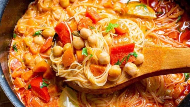 A close-up of a wooden spoon lifting vegetable and chickpea spaghetti in tomato sauce from a pot.