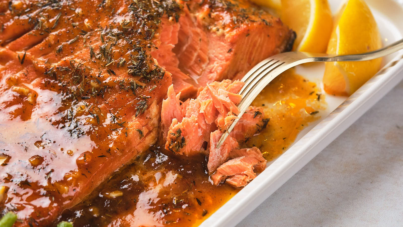 A fork flaking a piece of baked salmon covered in herb sauce with lemon wedges on the side.