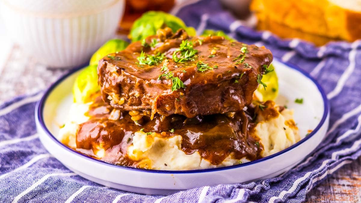 A plate of Salisbury steak with brown gravy served over mashed potatoes, garnished with chopped parsley, with Brussels sprouts on the side.