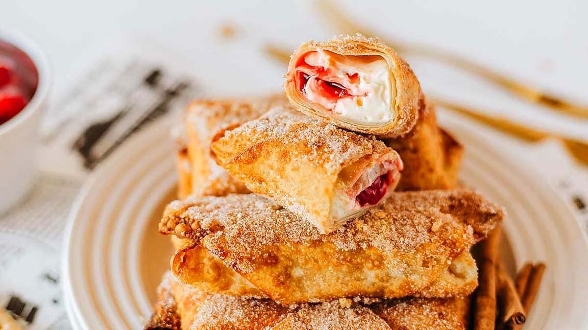 A plate of golden-brown, sugar-coated dessert chimichangas with a creamy and cherry filling, one cut open to show the inside.