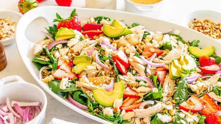 A large white dish with spinach salad topped with sliced strawberries, avocado, chicken, red onion, feta cheese, and sliced almonds. Small bowls with salad ingredients surround the dish.