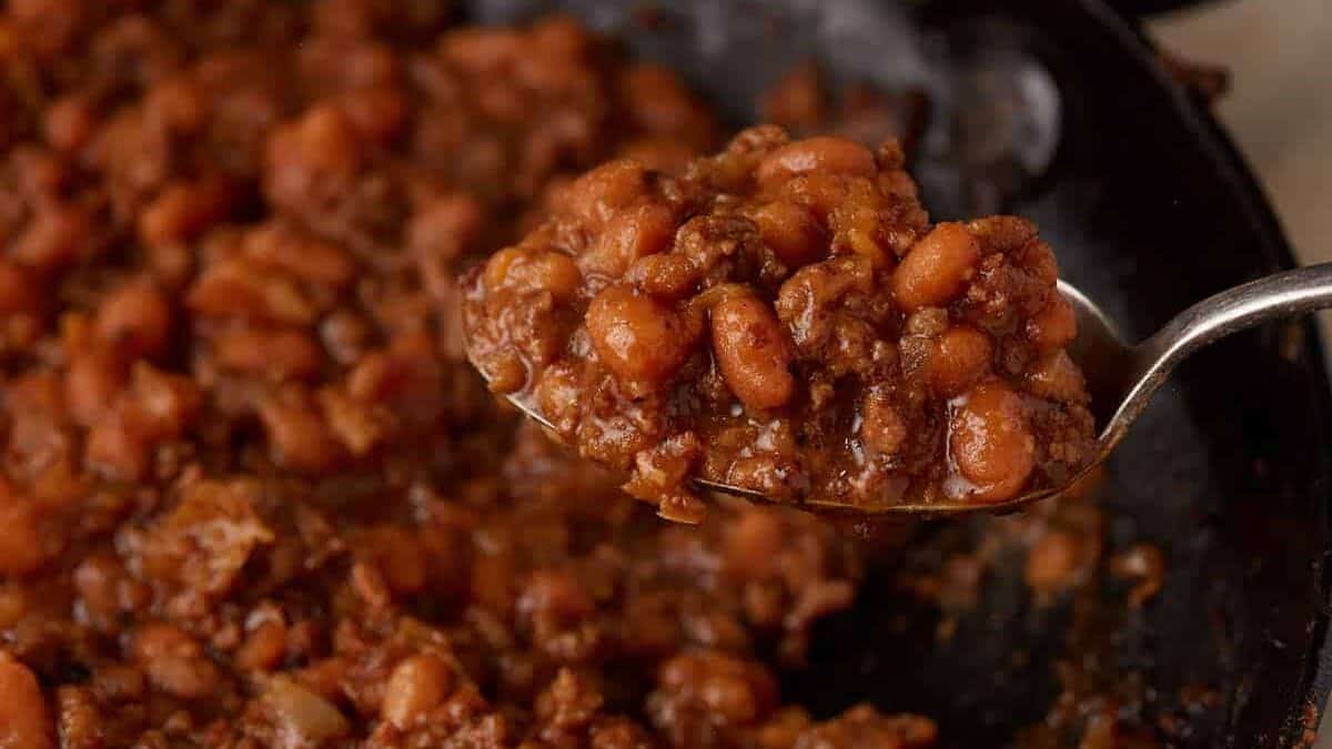 A close-up of a spoonful of baked beans with ground meat in a thick sauce, held over a pan filled with the same mixture.