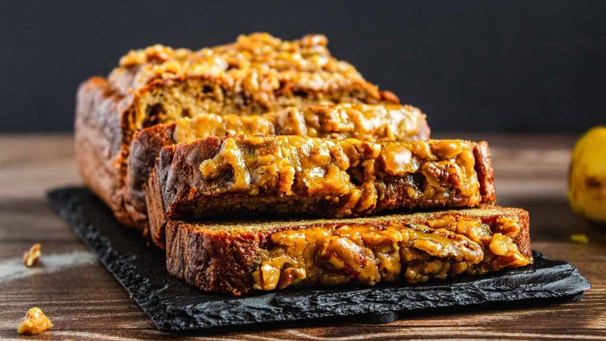 A loaf of banana bread topped with walnuts, partially sliced, displayed on a black slate board on a wooden surface.