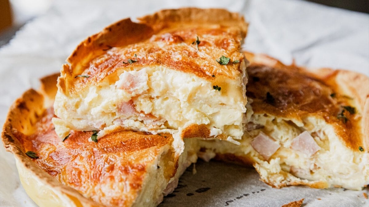 A close-up of a sliced quiche displaying a flaky crust, creamy filling with bits of ham, and melted cheese on top, garnished with chopped herbs.
