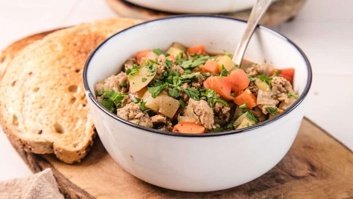 A bowl of hearty stew with ground meat, carrots, potatoes, and fresh herbs, with a spoon and bread on the side.