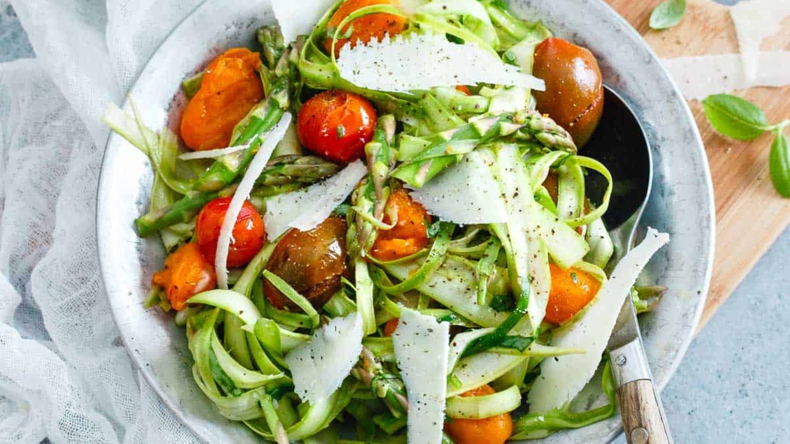 A bowl of shaved asparagus salad with cherry tomatoes and parmesan cheese shavings, garnished with black pepper.