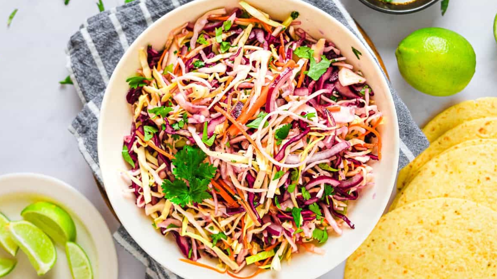 A bowl of colorful coleslaw with shredded cabbage, carrots, and cilantro, placed on a striped towel beside lime wedges and tortillas.