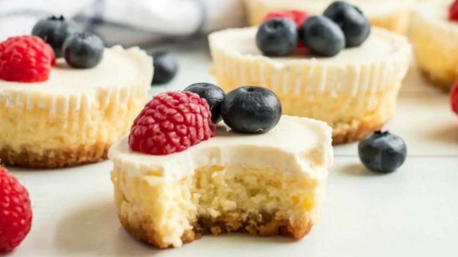 Mini cheesecakes topped with fresh blueberries and raspberries, one with a bite taken out, sit on a white surface.