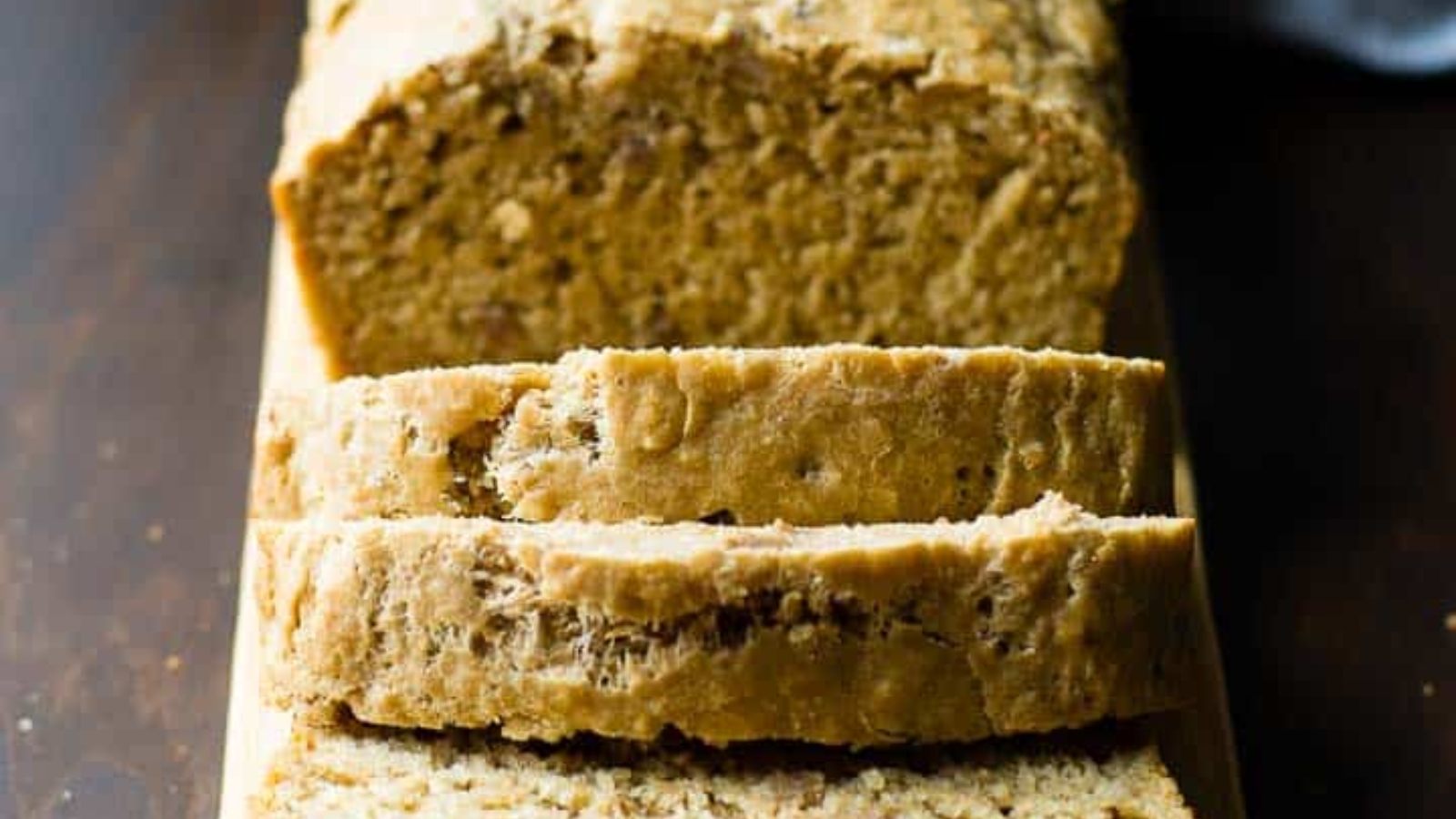 A loaf of bread on a wooden surface, partially sliced with three slices in front of the remaining loaf.