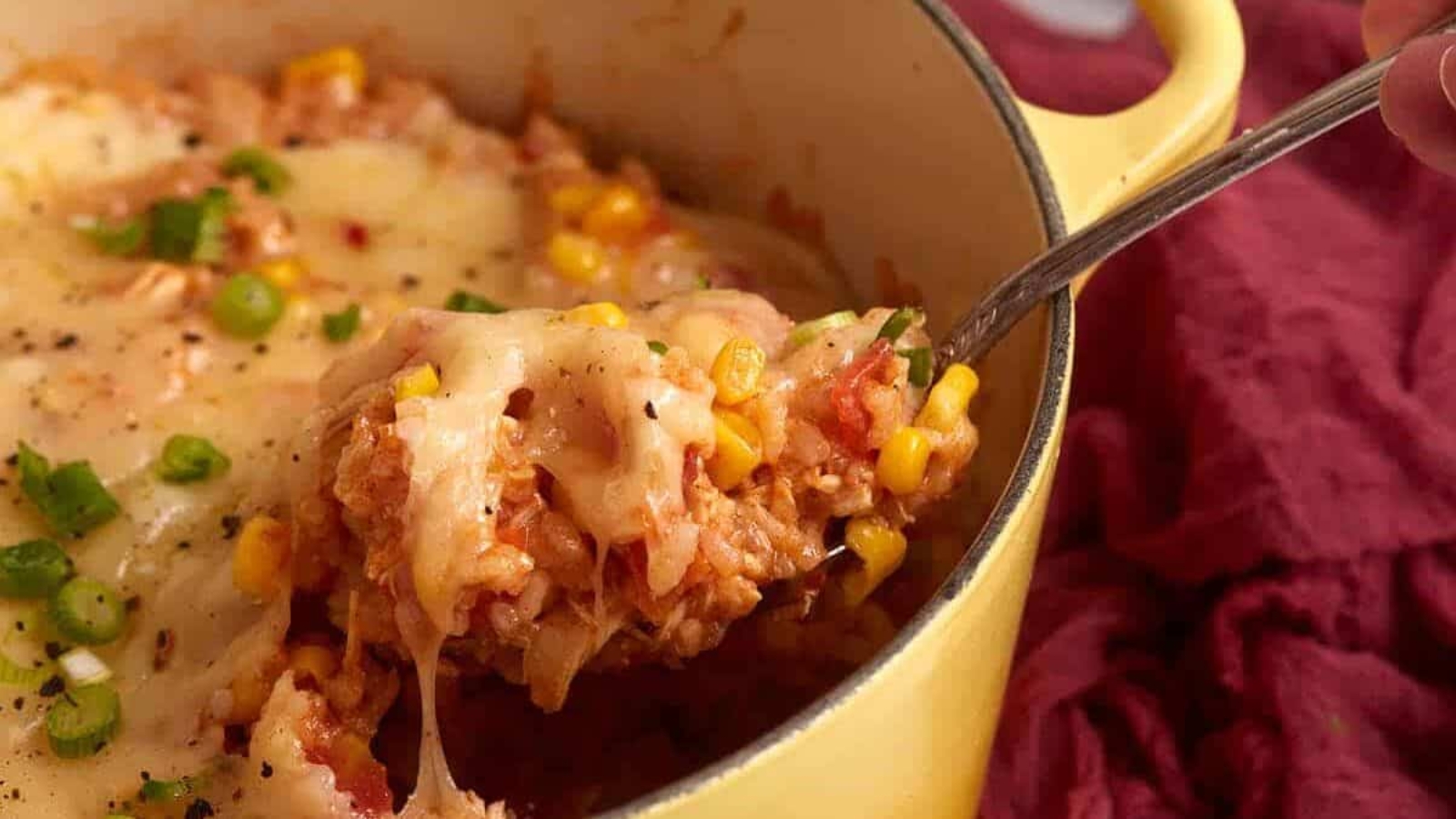 A spoon scoops cheesy rice, corn, tomatoes, and green onions from a yellow casserole dish.
