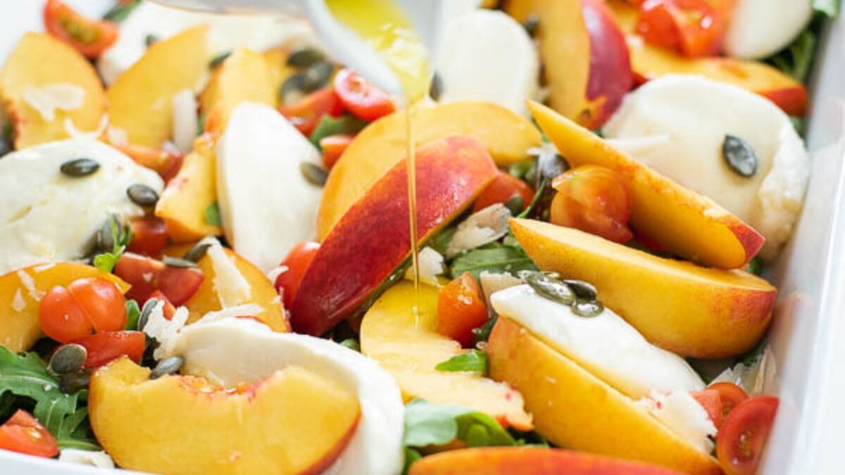 Sliced peaches, mozzarella, cherry tomatoes, and pumpkin seeds on a bed of greens with olive oil being drizzled on top.
