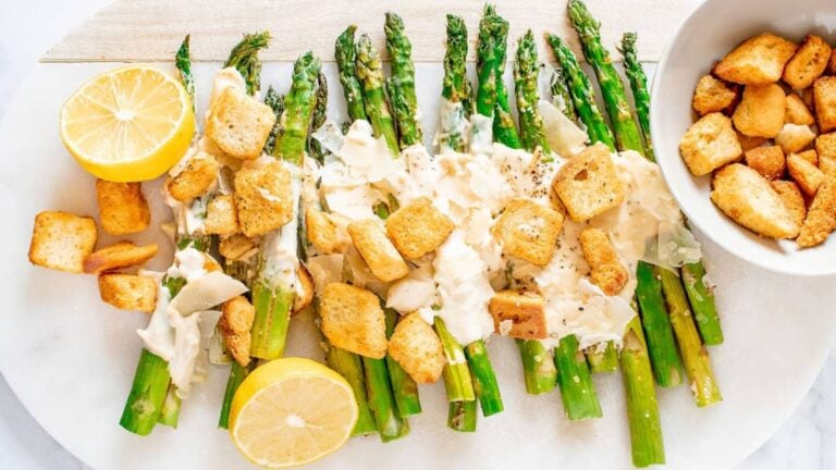 Grilled asparagus spears topped with creamy sauce, croutons, grated cheese, and black pepper, served with lemon halves on a white plate.