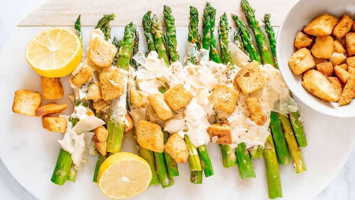 Grilled asparagus spears topped with creamy sauce, croutons, grated cheese, and black pepper, served with lemon halves on a white plate.