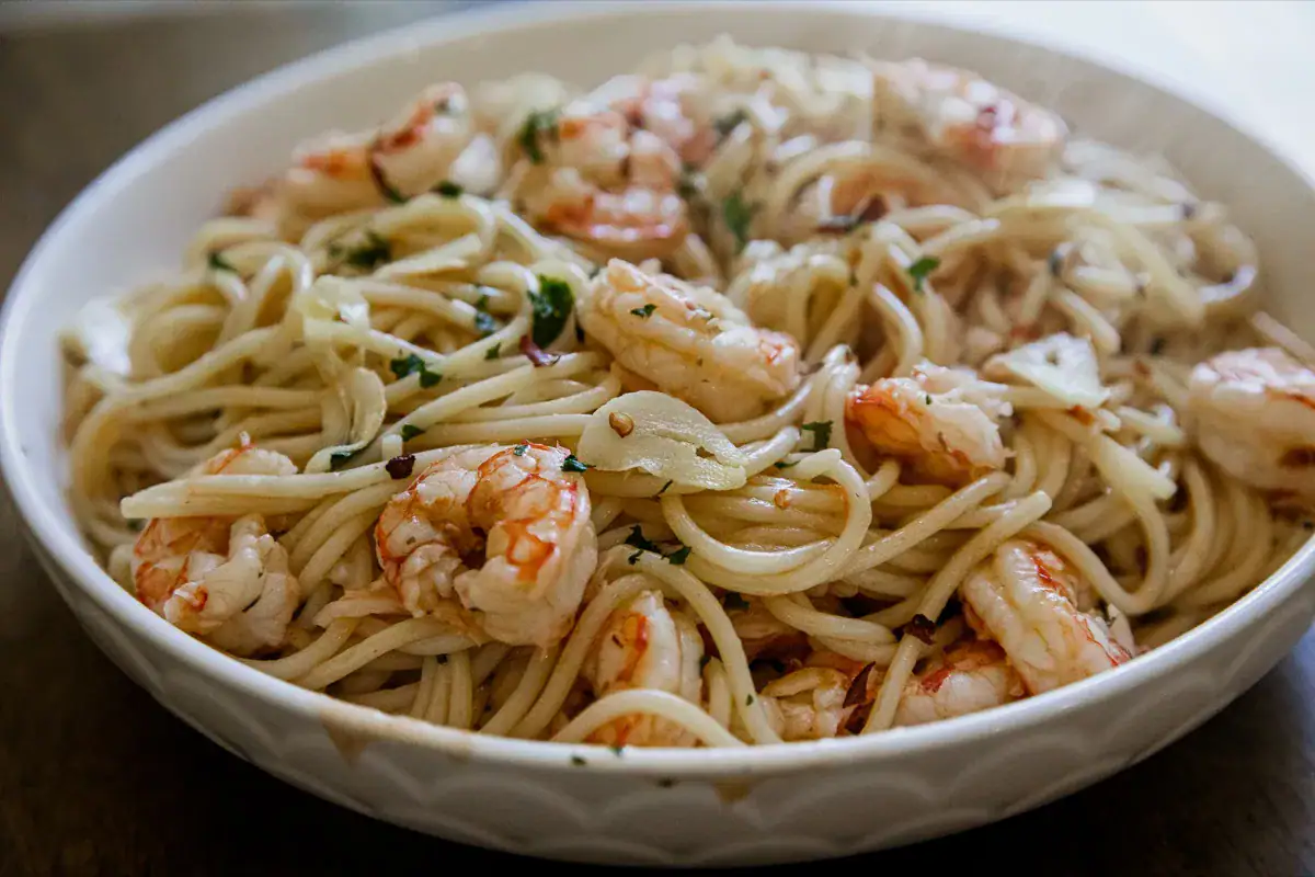 A white bowl filled with spaghetti pasta, shrimp, sliced garlic, and herbs, placed on a dark wooden surface.