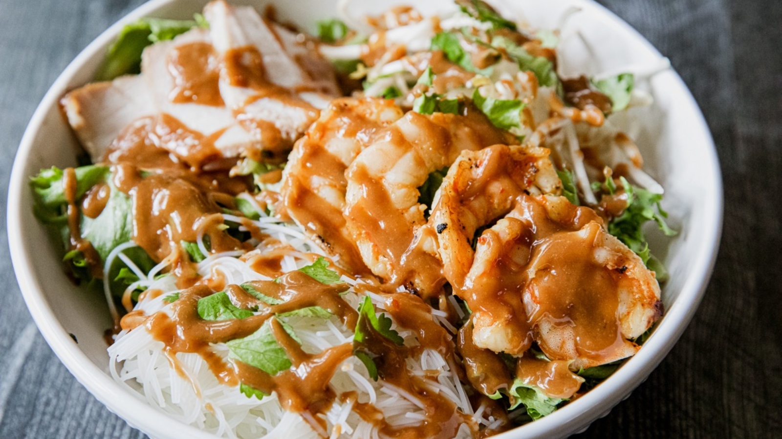 A bowl of salad with sliced chicken, shrimp, lettuce, rice noodles, and a generous drizzle of brown peanut sauce.