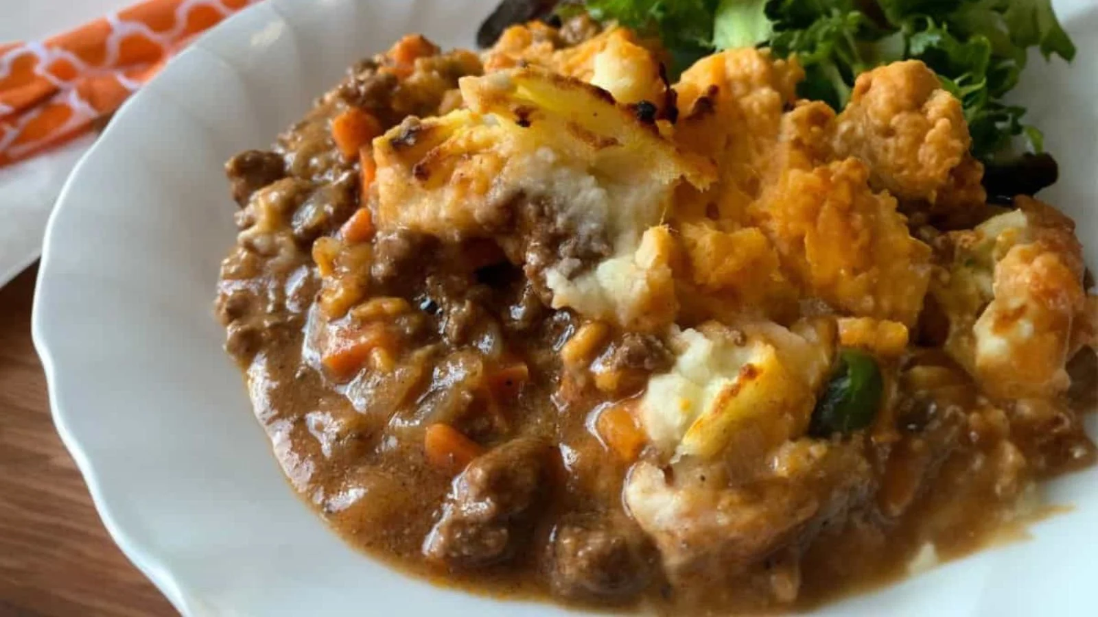 A close-up of shepherd's pie on a white plate, featuring ground meat, vegetables, and mashed potato topping, served with a side of leafy greens.