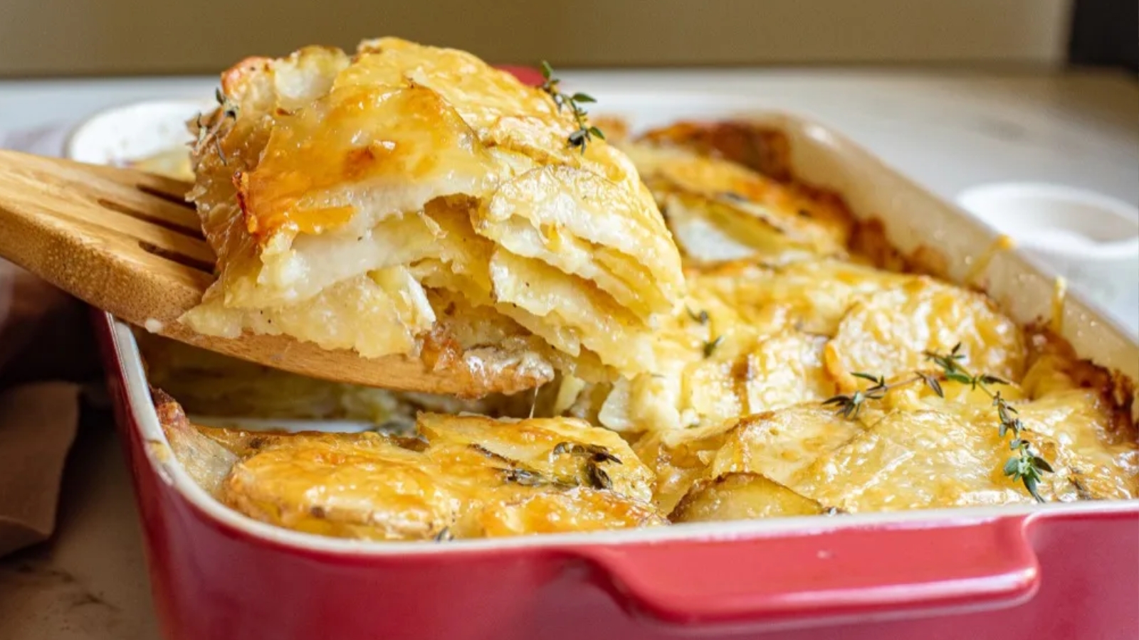 A close-up of a baked potato gratin in a red dish, with a serving being lifted out using a wooden spatula.