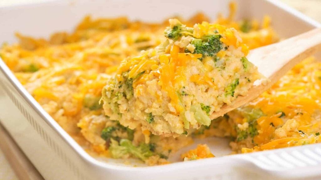 A wooden spatula lifts a serving of cheesy broccoli and rice casserole from a white baking dish.