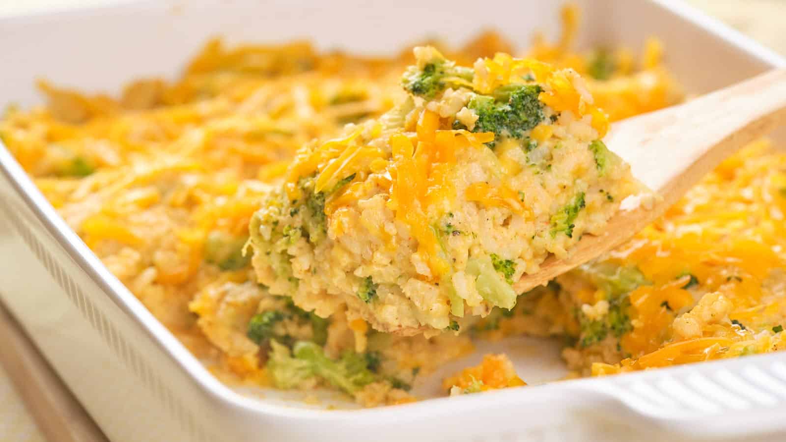 A wooden spatula lifts a serving of cheesy broccoli and rice casserole from a white baking dish.