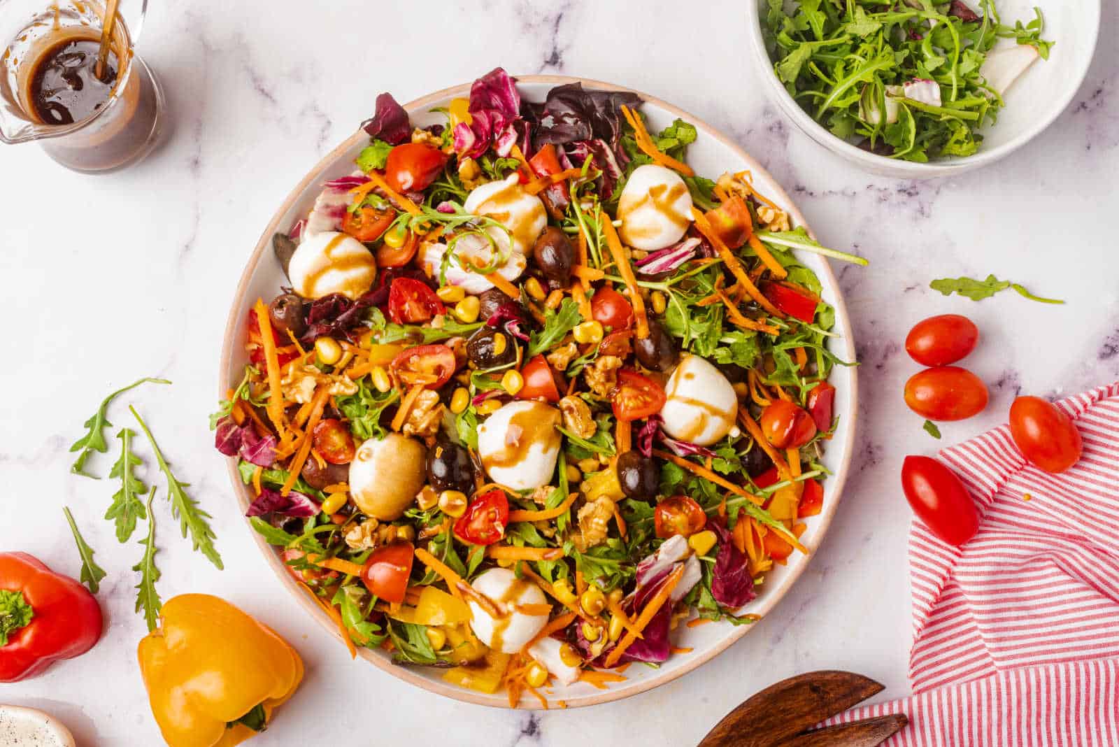 A colorful mixed salad with cherry tomatoes, lettuce, shredded carrots, olives, mozzarella balls, corn, and dressing on a plate, surrounded by salad ingredients and a bowl of greens.