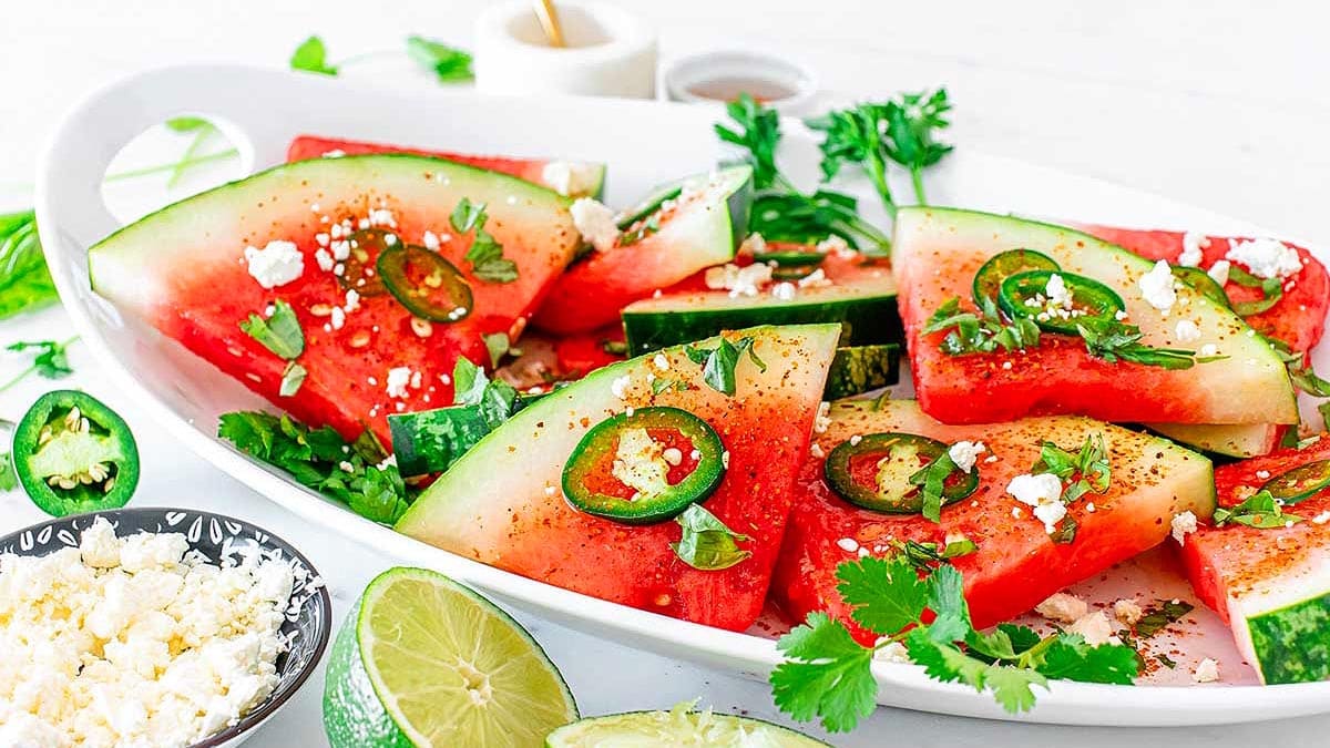 Sliced watermelon on a white platter topped with jalape&ntilde;o slices, crumbled cheese, herbs, and spices, with lime halves and cheese in bowls nearby.
