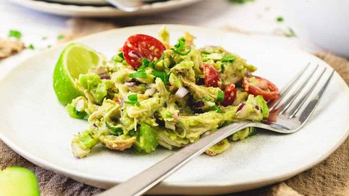 Chicken salad with cherry tomatoes, red onion, and lime wedge on a white plate with a fork.